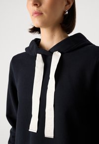 Jūrinės jūrų švarkelis su gobtuvu ir baltais firminiais raišteliais. Puikiai lygus tekstūra ir laisvas siluetas užtikrina komfortą.