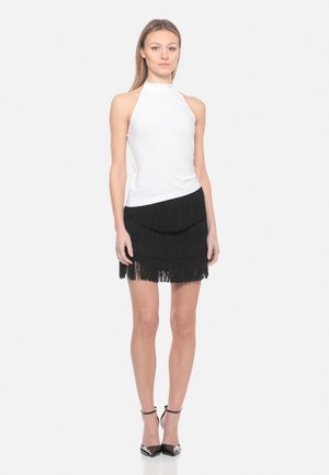 Giovane donna in piedi che indossa un top bianco smanicato con collo halter, minigonna nera a balze con frange e tacchi neri con cinturino alla caviglia su sfondo bianco.