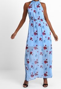Kvinna i en ärmlös ljusblå maxiklänning med röd och blå blommönster, halterneck, åtsittande midja, kombinerad med svarta klackskor.