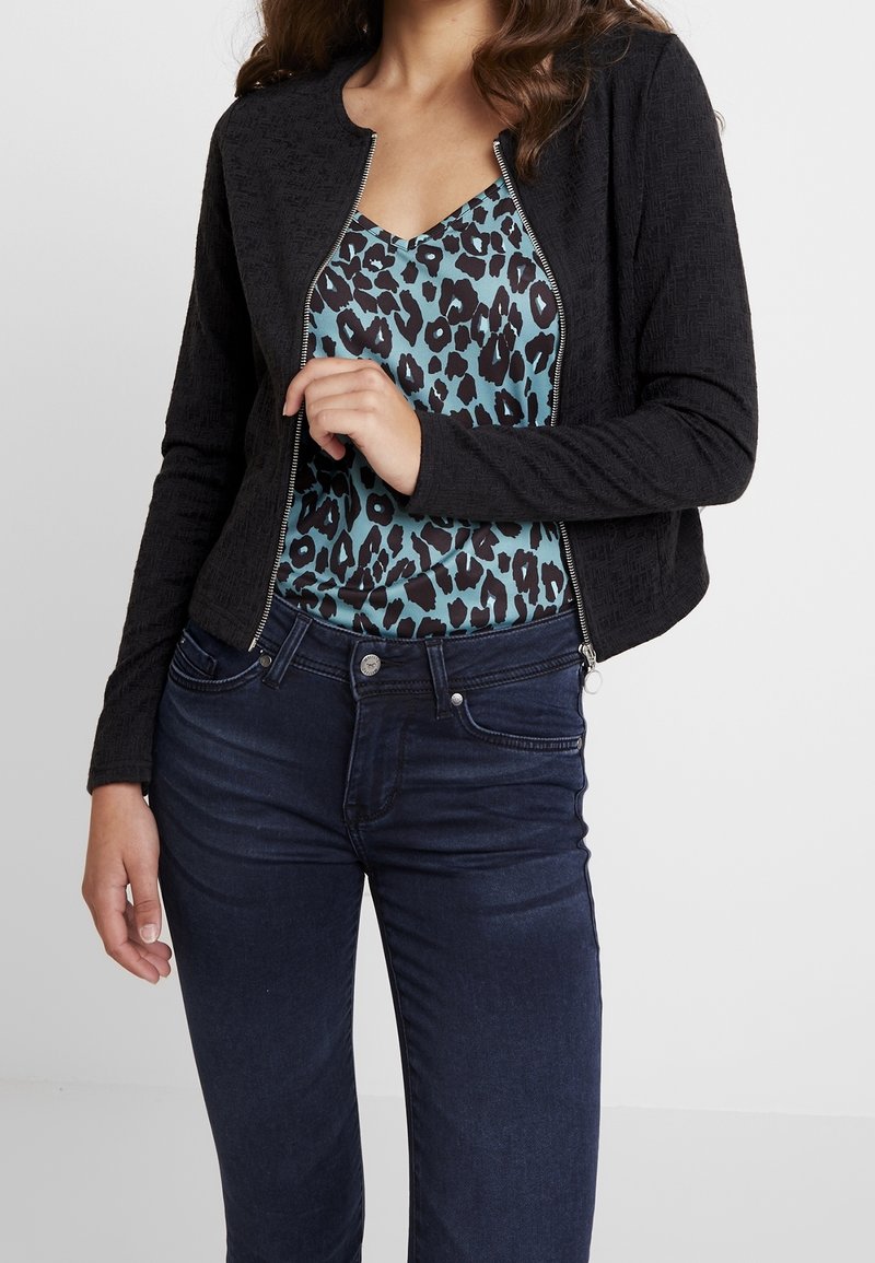 Černá texturovaná bunda s kulatým výstřihem, zapínáním na zip, ve spojení s tyrkysovým topem s leopardím potiskem a tmavě modrými úzkými džíny.