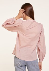 Blusa de color rosa claro con mangas abullonadas, superficie texturizada con pequeños puntos en relieve y un escote redondo. Llevada con jeans de cintura alta.
