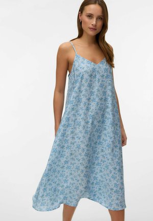 Femme portant une robe midi sans manches à fleurs, bleu clair, avec de fines bretelles, debout devant un fond blanc uni.
