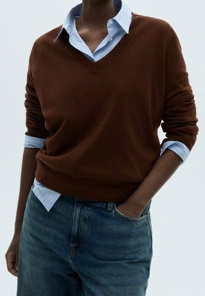 Personne portant un pull marron à col en V sur une chemise à rayures bleues avec manches retroussées et un jean bleu, une main dans la poche.