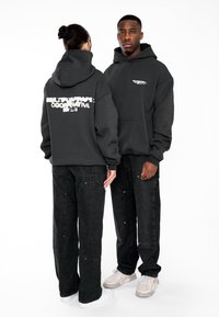 Schwarze Kapuzenjacke mit weißem Grafiktext auf der Rückseite und Vorderseite, kombiniert mit dunklen Jeans. Beide Kleidungsstücke haben eine lockere, übergroße Passform.