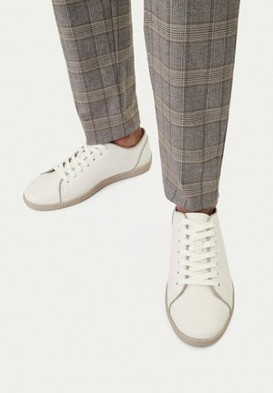 Weiße Low-Top-Sneaker mit beigefarbenen Sohlen, getragen zu grauen karierten Hosen vor schlichtem weißem Hintergrund.