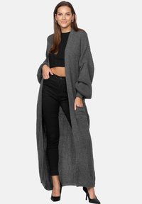 Cardigan lungo grigio con texture rigata, apertura frontale, maniche oversize e due tasche frontali, indossato sopra un crop top nero e pantaloni neri aderenti.