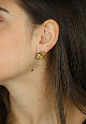 Orecchio sinistro di una donna con un orecchino a perno dorato a forma di nodo e una piccola lentiggine scura sul collo sotto l'orecchio.