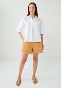 Chemise blanche à manches courtes avec broderie florale dorée, associée à un short beige et des tongs marron. Design simple, style décontracté.