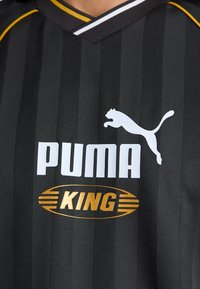 Juodas dryžuotas sportinis džersis su baltu "PUMA" logotipu ir auksiniu tekstu "KING". Dėmesys skiriamas viršutinei krūtinės daliai, rodant audinio tekstūrą ir detales.