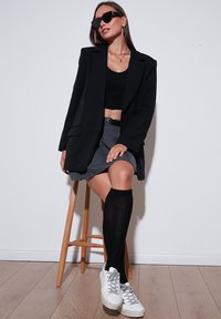 Blazer negru supradimensionat, top negru scurt, fustă gri, ciorapi negri până la genunchi și adidași albi, așezată pe un taburet de lemn, pe un fundal simplu.
