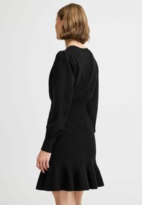 Svart stickad klänning med uppsvällda långa ärmar, en figurnära bodice och en utsvängd fåll. Slät textur; minimal detaljering.