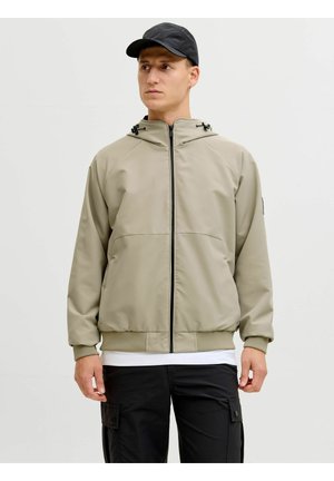 Homme portant une veste à capuche zippée beige clair, un pantalon cargo noir et une casquette noire, debout contre un fond clair uni, regardant vers la droite.