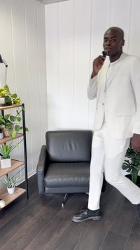 Blazer e pantaloni bianchi indossati con una camicia bianca, mentre si cammina vicino a una poltrona in pelle nera e a una libreria in legno con piante. Pavimento in legno scuro.