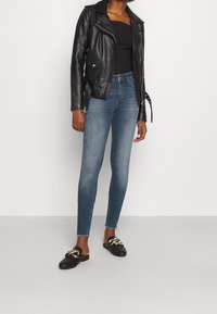 Chaqueta de moto de cuero negra, camiseta ajustada negra, vaqueros skinny azules, zapatos negros sin cordones con detalle de cadena dorada. Diseño simple y moderno.