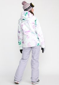 Skijacket met een witte basis, roze en groene bloempatroon, aansluitende capuchon. Gepaard met lichtgrijze skibroek en zwarte handschoenen.