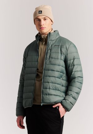 Junger Mann mit beige-farbenem Beanie, grüner Daunenjacke und braunem Reißverschluss-Pullover, steht mit einer Hand in der Tasche vor einem schlichten Hintergrund.
