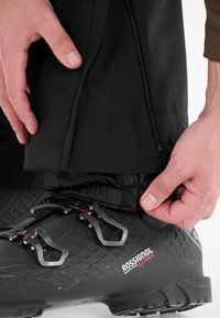 Mustat hiihtosaappaat, joissa on teksturoitu pinta ja kolme metallista solkea. Käsi säätämässä mustien housujen säärtä, joissa on vetoketjullinen sivuaukko.