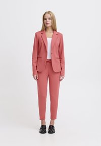 Roze blazer met twee voorzakken, gecombineerd met bijpassende tapered broek. Gladde stof, slim fit, afgewerkt met zwarte schoenen.