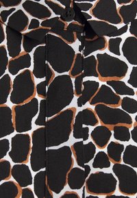 Sort og hvid bluse med giraffemønster, der har uregelmæssige brune pletter og en knaplukning i kraven. Blød stofstruktur.