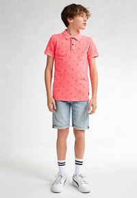 Kirkkaanpunainen poolopaita, jossa on pieniä tummansinisiä kuvioita, yhdistettynä käännettyihin farkkushortseihin. Valkoiset lenkkarit ja raidalliset sukat täydentävät asun.