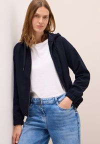 Mörkblå zip-up sweatshirt med huva, ribbade ärmslut och framfickor, parat med en vit t-shirt och ljusblå denimjeans.