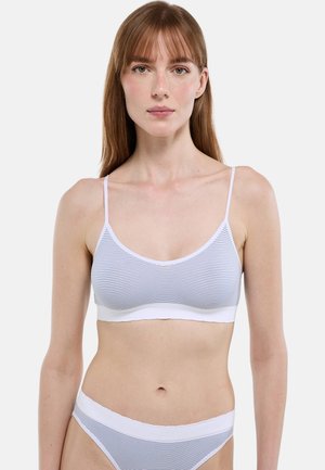 Jeune femme aux cheveux châtains clairs et raides portant un soutien-gorge blanc à fines rayures bleues et une culotte assortie, sur un fond uni.