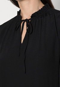 Svart blus med plisserad krage och nyckelhål fram, med justerbara band för stängning, tillverkad av ett mjukt, strukturerat tyg.