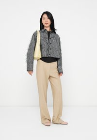 Jonge vrouw staat in een gestructureerde grijze cropped jack, beige wijde broek met knopen bij de manchetten, lichtroze schoenen en een crèmekleurige schoudertas.
