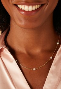 Gouden ketting met kleine witte kralen, gedragen over een roze satijnen top. Heeft een delicate uitstraling, gladde textuur en subtiele glans.