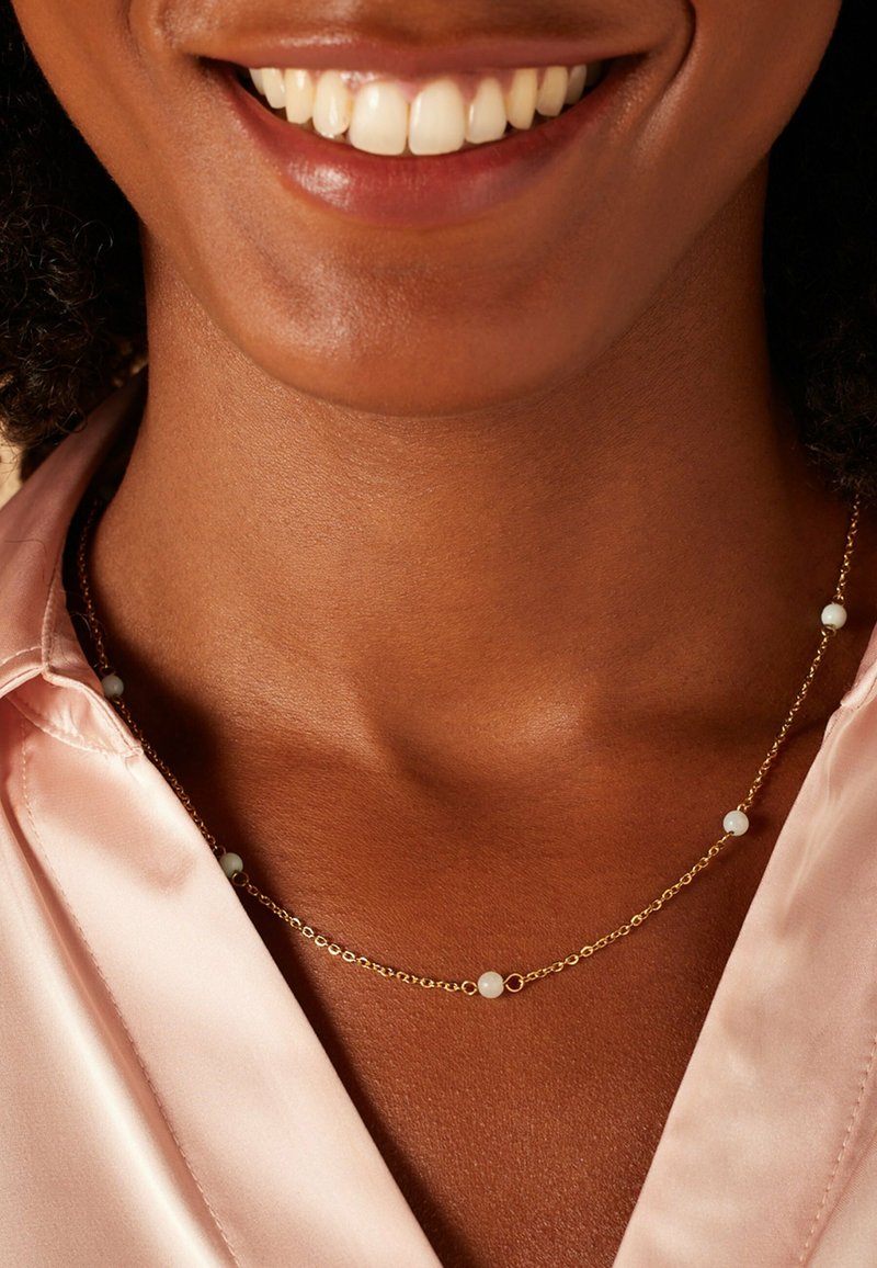 Gouden ketting met kleine witte kralen, gedragen over een roze satijnen top. Heeft een delicate uitstraling, gladde textuur en subtiele glans.