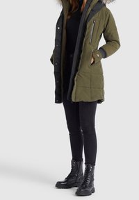 Olivgrön vadderad jacka med svarta detaljer, huva kantad med fuskpäls, tryck- och dragkedjestängningar samt sidofickor. Buren över svarta smala jeans och stövlar.