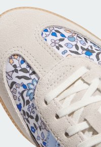 Baskets en daim beige avec des panneaux en tissu floral, des lacets blancs et une semelle en caoutchouc texturée. Accents bleu et orange sur le motif floral.