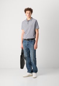 Camisa polo gris con acentos amarillos, jeans azules y zapatillas blancas. Sosteniendo una mochila negra, de pie en un fondo neutro.