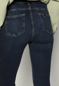 Jeans de denim azul oscuro con cinco bolsillos, costuras visibles y un ajuste ceñido. Material de algodón con ligera textura, con costuras en contraste.