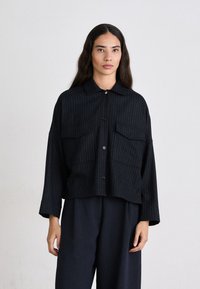 Schwarz gestreiftes Hemd mit Knöpfen und Kragen, zwei vorderen Taschen und cropped Länge. Das Material wirkt strukturiert. Kombiniert mit dunklen Hosen.