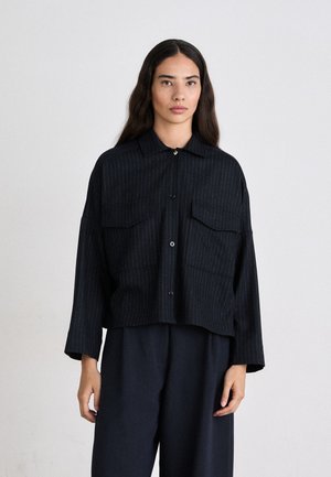 Camicia a righe nere con colletto, due tasche frontali e lunghezza cropped. Il tessuto sembra texturizzato. Abbinata a pantaloni scuri.