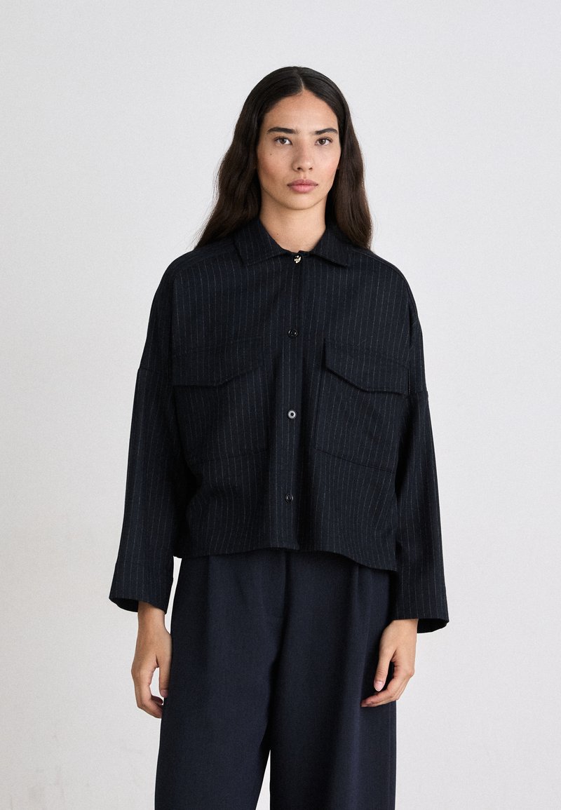Schwarz gestreiftes Hemd mit Knöpfen und Kragen, zwei vorderen Taschen und cropped Länge. Das Material wirkt strukturiert. Kombiniert mit dunklen Hosen.