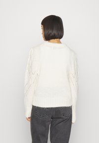 Fetziges weißes Sweatshirt mit strukturiertem Strick, das einen gerippten Saum und offene Maschenmuster an den Ärmeln aufweist. Getragen mit dunklen grauen Hosen.