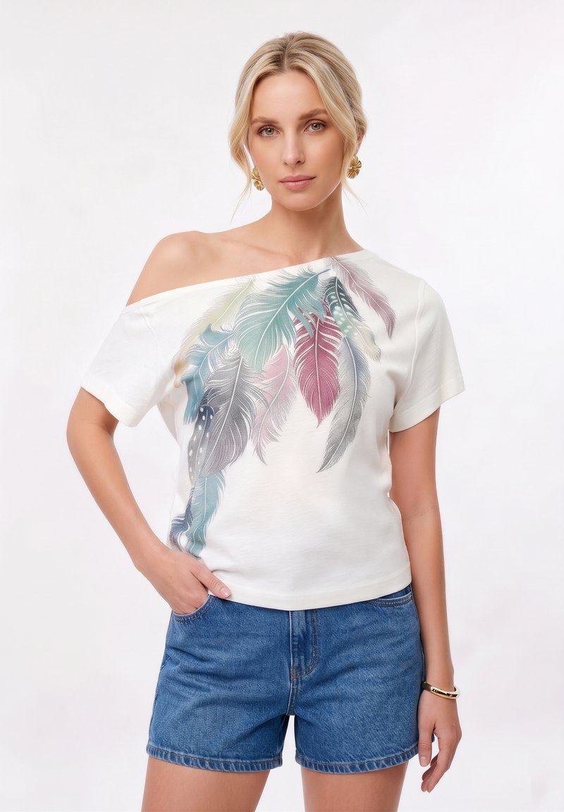 Femme blonde portant un T-shirt blanc épaules dénudées avec un imprimé plumes multicolores, un short en jean bleu, ainsi que des boucles d'oreilles et un bracelet dorés.