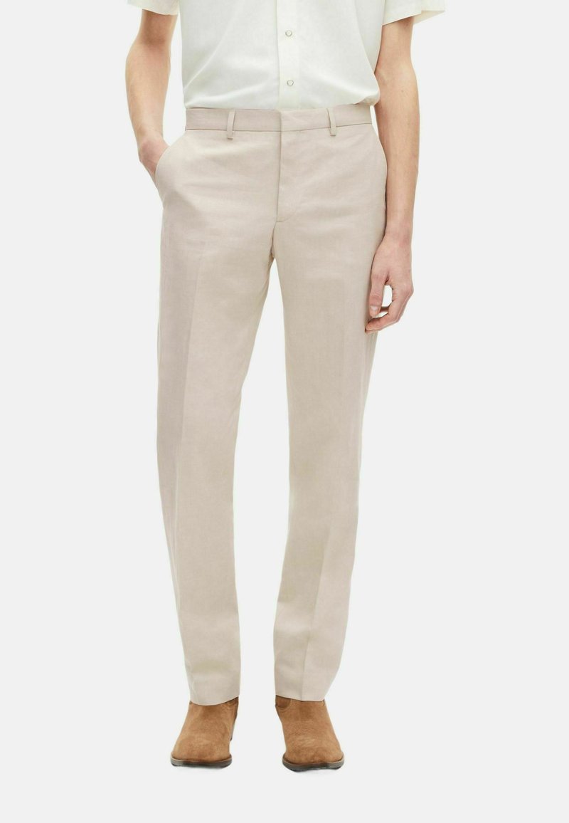 Helle beige Leinenhosen mit geradem Schnitt. Sie verfügen über zwei Vordertaschen und eine flache Vorderseite und werden mit braunen Wildleder-Stiefeletten kombiniert.