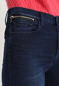 Jeans oscuros de corte slim con una textura suave, que presentan un bolsillo frontal con cremallera y un diseño clásico de cinco bolsillos.
