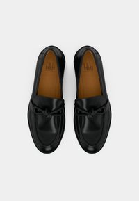 Μαύρα δερμάτινα μοκασίνια με λεία υφή, στρογγυλεμένη μύτη και διακοσμητικό κόμπο μπροστά. Δερμάτινη εσωτερική σόλα και ελάχιστα ραφές ως διακοσμητικό στοιχείο.