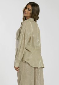 Camisa de botones metálica beige con mangas largas y un corte relajado, que presenta un brillo sutil y un bolsillo en la parte izquierda.