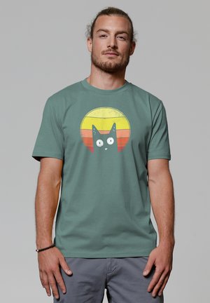 Groene katoenen t-shirt met een grafisch ontwerp van een zwarte kat binnen een cirkelontwerp van een vintage zon met gele en oranje horizontale strepen.