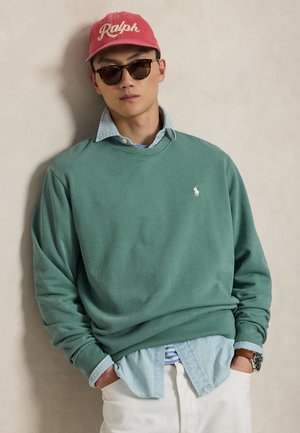 Homme portant une casquette rouge "Ralph", des lunettes de soleil, un sweat-shirt vert sur une chemise bleue boutonnée, un pantalon blanc et une montre-bracelet, les mains dans les poches.