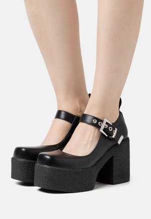 Black patent leather platform shoes with a textured rubber sole, round toe, and ankle strap featuring a silver buckle detail.