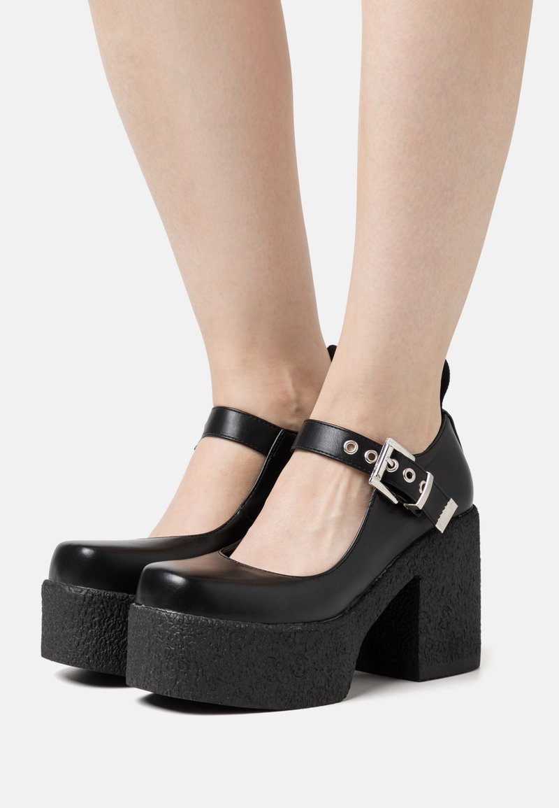 Black patent leather platform shoes with a textured rubber sole, round toe, and ankle strap featuring a silver buckle detail.