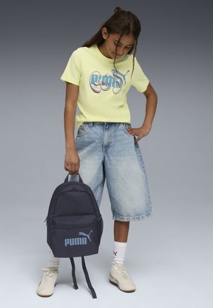 Geel grafisch T-shirt met blauw logo, lichtblauwe denim shorts, donkerblauwe rugzak met Puma-logo en witte sneakers met zwarte accenten.