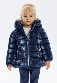 Marinblå puffjacka med glänsande yta, horisontellt kviltad design, huva, dragkedjestängning och sidofickor. Bärs över en ljus topp och jeans.