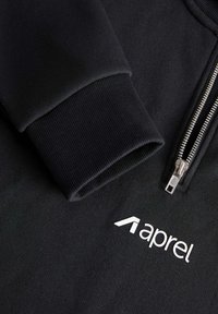 Sudadera negra con cremallera y puños acanalados, con una textura suave y un logo blanco "aprel" impreso en el frente.